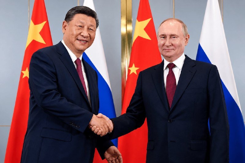 Two men in dark suits shake hands, smiling at the camera, with Chinese and Russian flags displayed behind them.