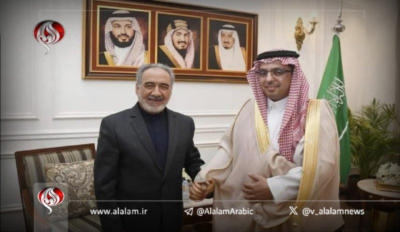 Two men shake hands in a formal room, one in a dark suit and the other in traditional Arab attire, with three framed portraits and a green flag visible behind them.