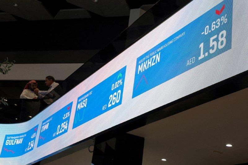 A digital stock ticker displays company listings including Gulf Navigation, Itthmaar, MASQ, and MKHZN with prices in AED, while two people converse on a balcony behind it.