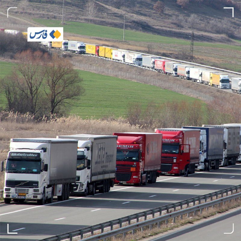 A long line of semi-trucks queues along a highway and an adjacent road, set against green fields and distant hills.