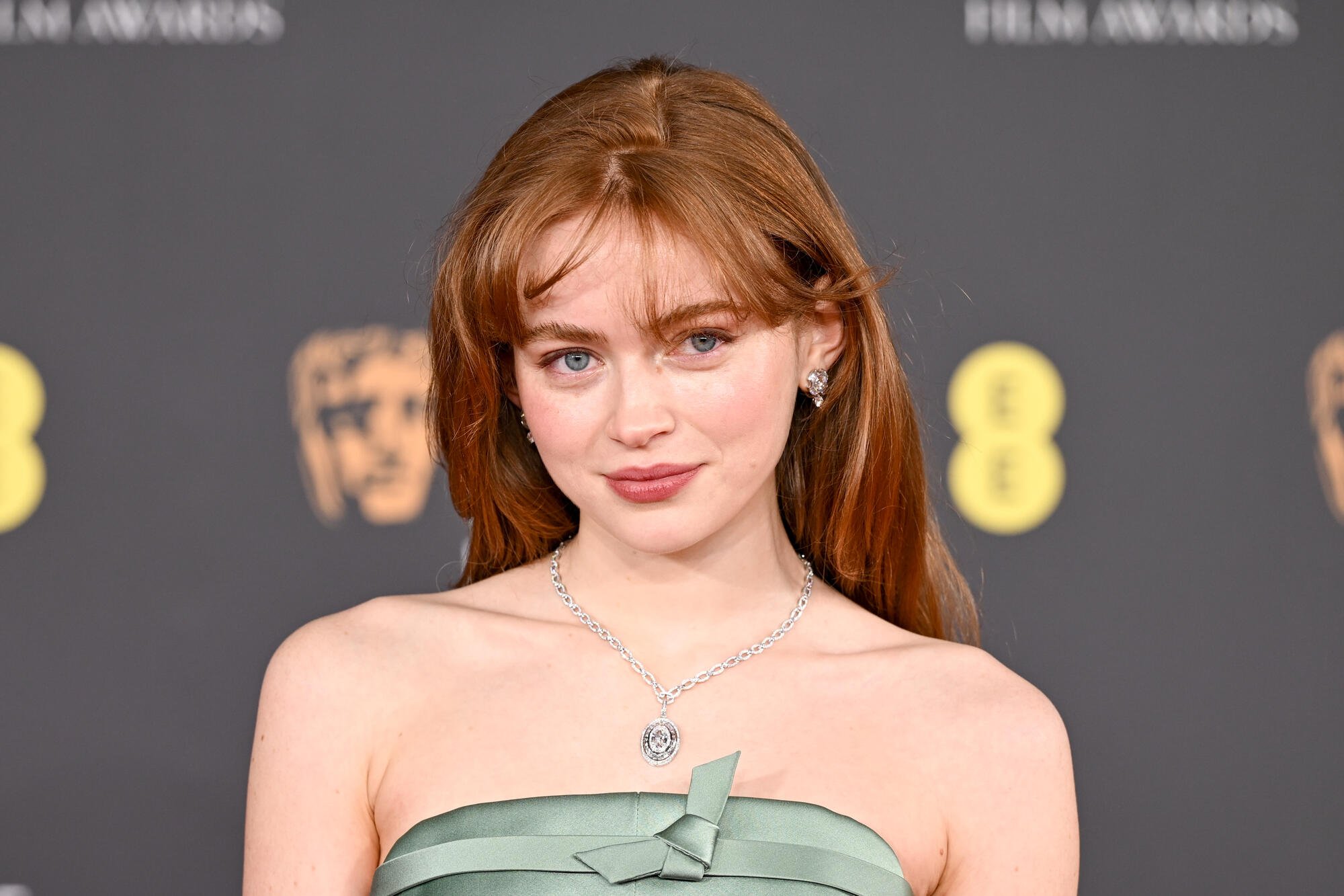 A woman with long auburn hair and bangs wears a green strapless dress and silver pendant necklace, standing before a dark backdrop displaying the BAFTA logo and the number "8."