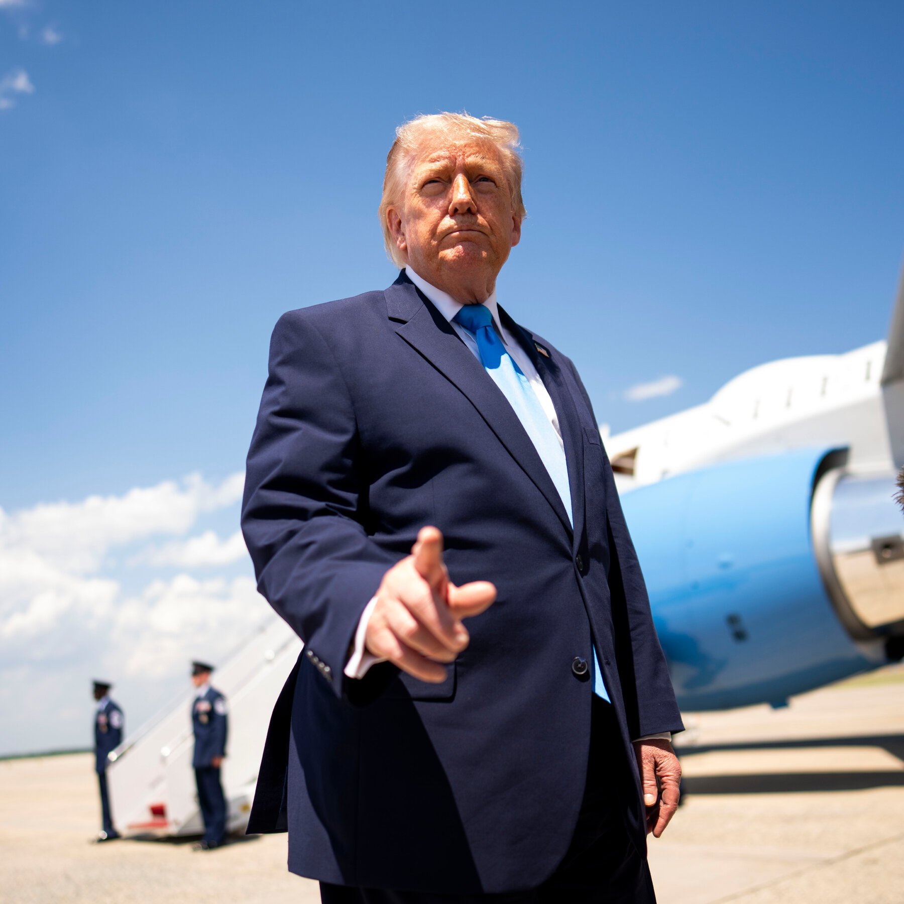 An older man with blonde hair in a navy suit and blue tie points toward the camera on a tarmac, with a blue and white aircraft and two uniformed personnel in the background.