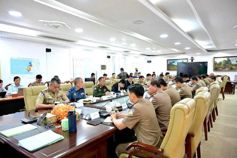 Officials in military and civilian uniforms sit around a long conference table in a bright meeting room with maps and a large screen visible on the walls.