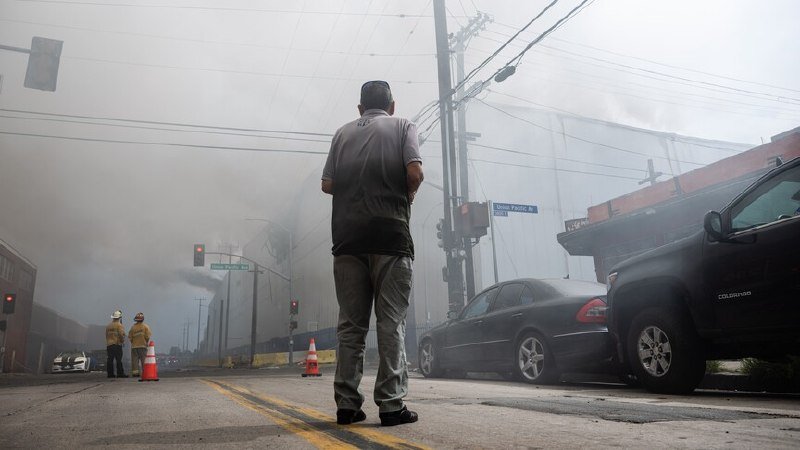 A man in a gray shirt stands on a smoke-filled street observing firefighters in yellow gear near a traffic cone, with parked cars and power lines visible.