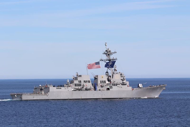 A gray U.S. Navy guided-missile destroyer marked with hull number 107 sails across open blue waters beneath a clear sky, flying an American flag.