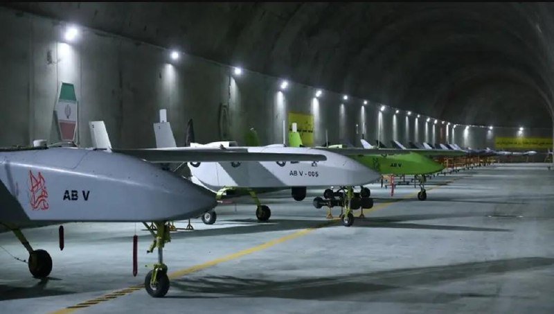 A row of unmanned aerial vehicles marked "AB V" and "AB V-005" sits parked inside a large, lit concrete hangar or tunnel.