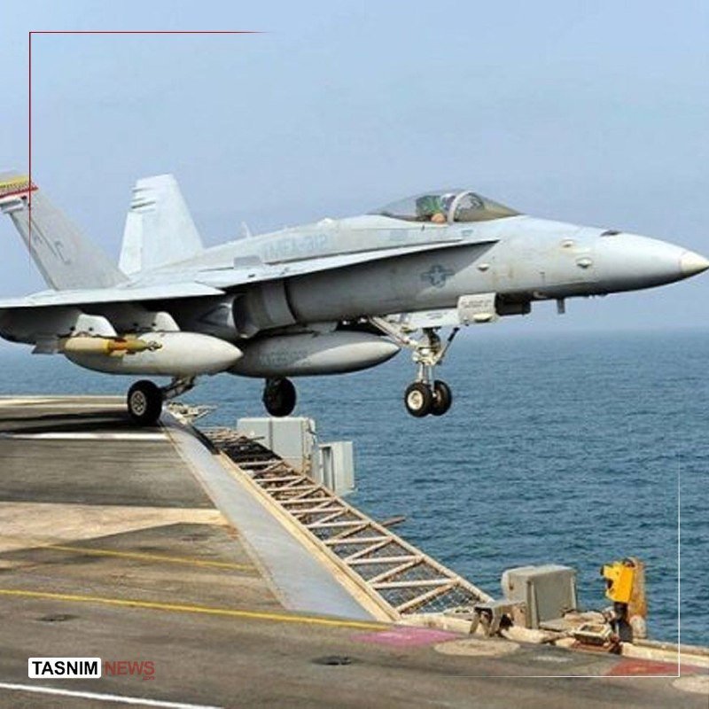 A gray fighter jet with extended landing gear launches off an aircraft carrier's ski-jump ramp over the ocean.