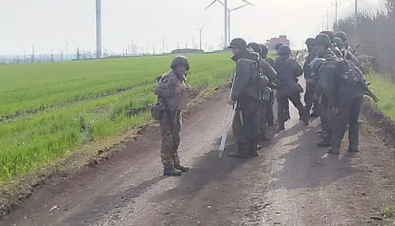Armed soldiers in camouflage uniforms and helmets stand gathered on a rural dirt road, with one facing a group holding rifles and gear, while wind turbines are visible in the distant green field.