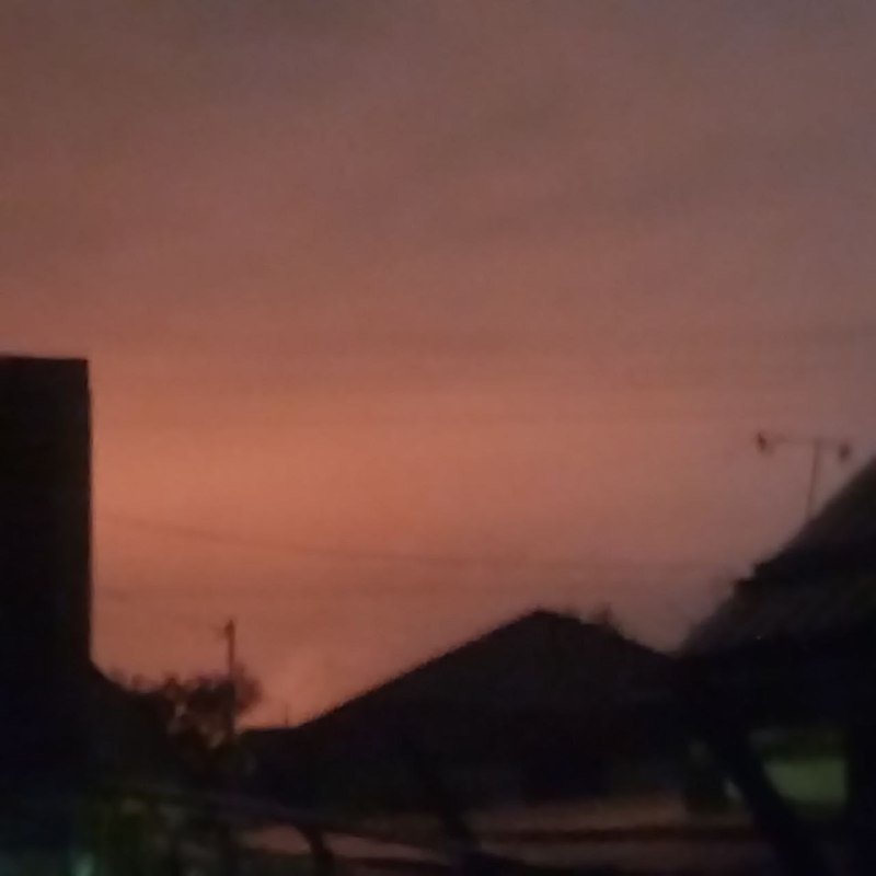 A hazy pink and orange sunset sky appears above silhouetted rooftops and power lines.