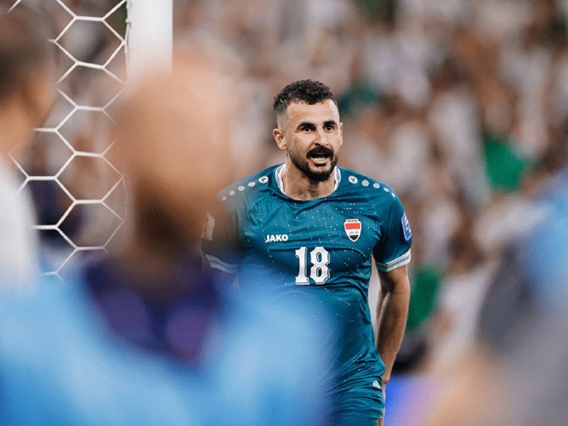 A bearded soccer player in a teal JAKO jersey numbered 18 with an Iraq emblem stands on the field, smiling, with a goal net and blurred crowd behind him.