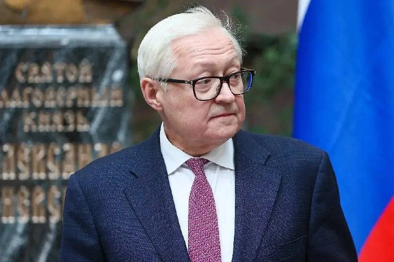 A white-haired man wearing glasses, a dark suit, and a pink patterned tie stands beside a Russian flag with a partially visible monument bearing Cyrillic text in the background.