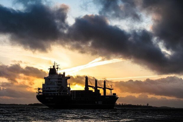 A large cargo ship with cranes sails across a calm sea at sunset, silhouetted against a sky filled with dark clouds and golden light.
