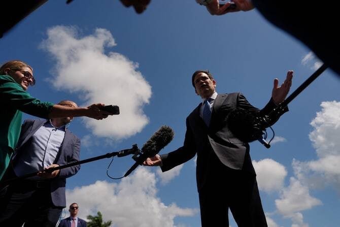 A man in a suit gestures while speaking to reporters holding microphones and recording devices outdoors under a cloudy blue sky.