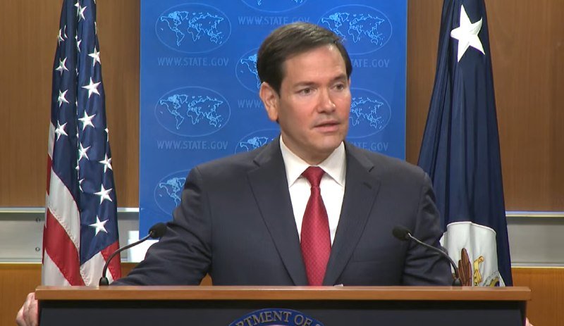 A man in a dark suit and red tie speaks at a podium bearing the Department of State seal, flanked by U.S. and Department of State flags against a blue State.gov backdrop.