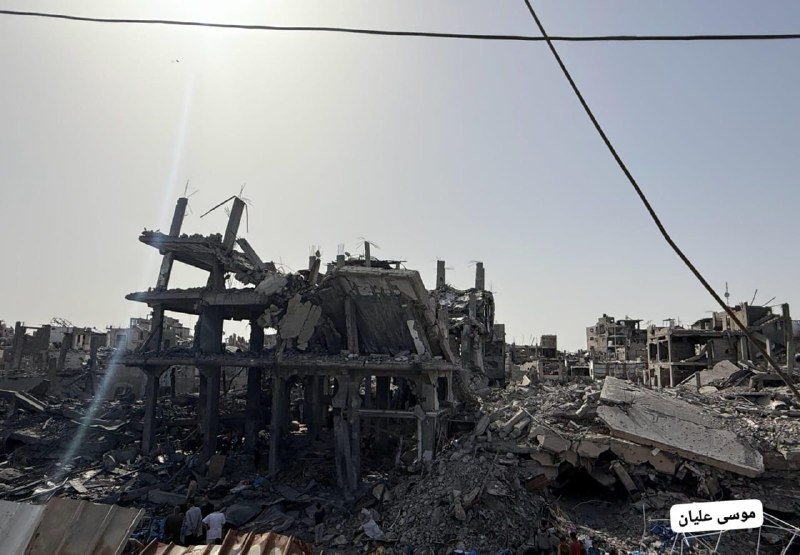 A partially collapsed concrete building stands amid extensive rubble and debris under a bright sky, with power lines crossing overhead.
