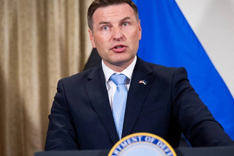 A man in a dark suit and blue tie speaks at a podium bearing a "Department of Defense" seal, with blue and black flags visible behind him.