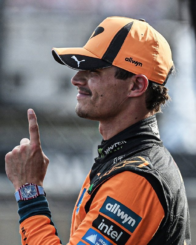 A smiling man in an orange and black racing suit with sponsor logos points upward, wearing a matching cap and a wristwatch.