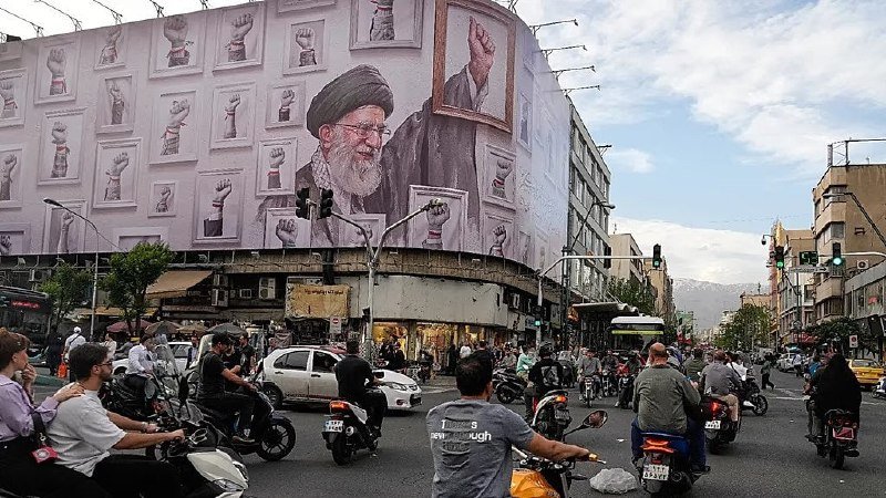 A large mural of a bearded cleric in a black turban surrounded by raised fists covers the side of a building above a busy street filled with motorcyclists, cars, and pedestrians.