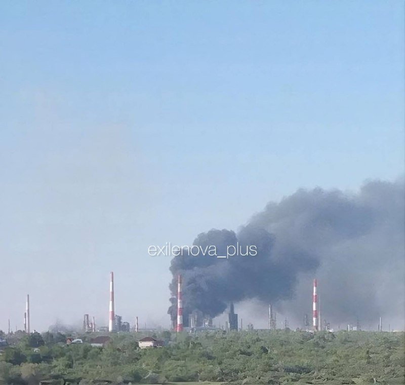 Thick black smoke rises from an industrial facility with tall red-and-white striped smokestacks, set against a clear sky with green trees in the foreground.