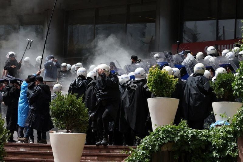Riot police in white helmets and dark uniforms form a line with shields and gas masks amid smoke outside a glass-fronted building, facing a crowd including a person holding up a phone.
