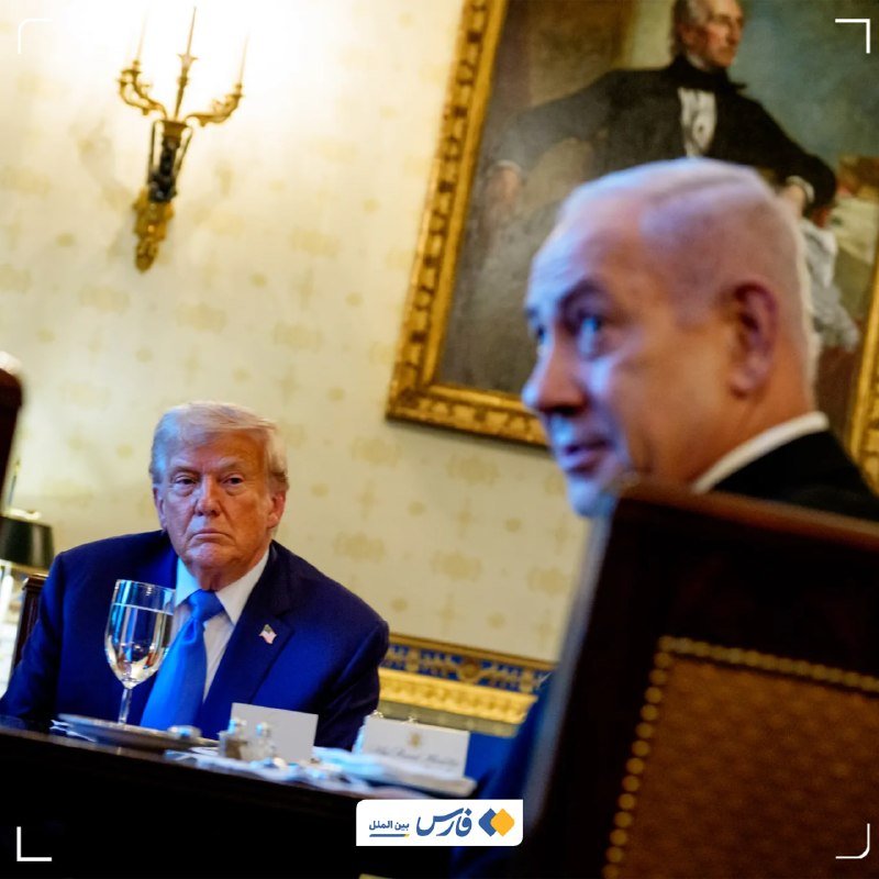 Two men in suits sit at a formal table in an ornately decorated room with a gold-framed portrait and wall sconce in the background.