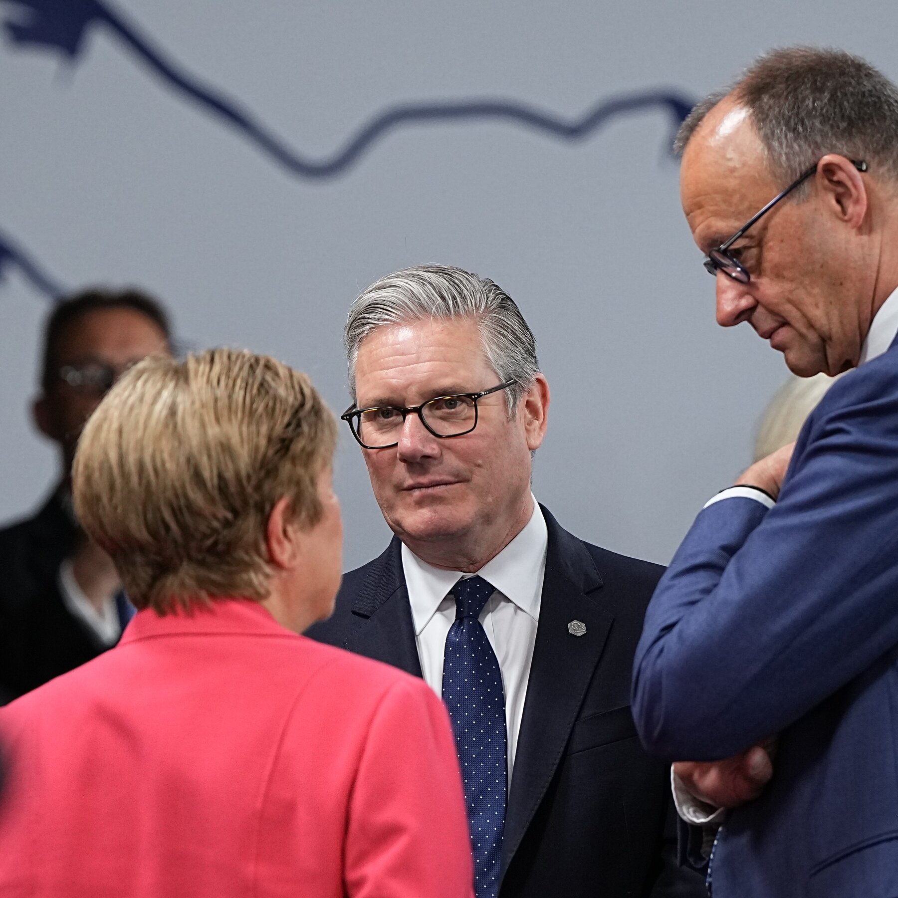A gray-haired man in a dark suit, white shirt, and dotted blue tie stands between a blonde woman in a pink jacket and another bespectacled man in a blue suit.