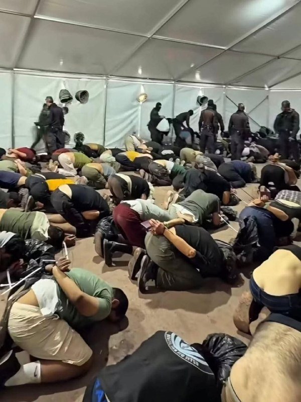 Numerous people kneel with heads down on the floor inside a large white tent, while uniformed officers stand along the perimeter walls.