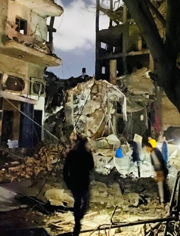 People walk through rubble and debris surrounding severely damaged, partially collapsed multi-story buildings at night.