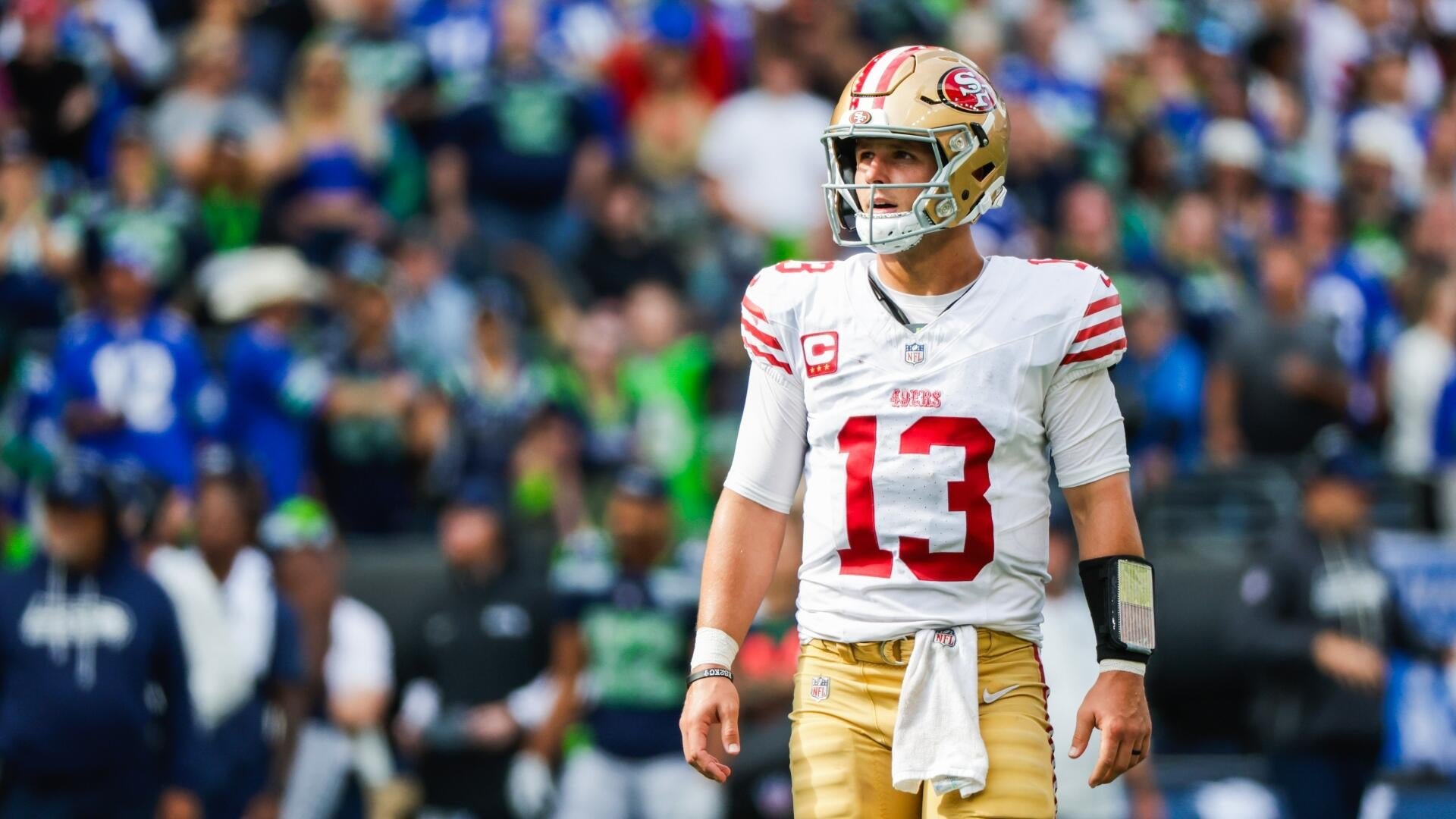 A football player wearing a white San Francisco 49ers jersey with the number 13 and a gold helmet stands on the field, with a blurred crowd visible in the background.