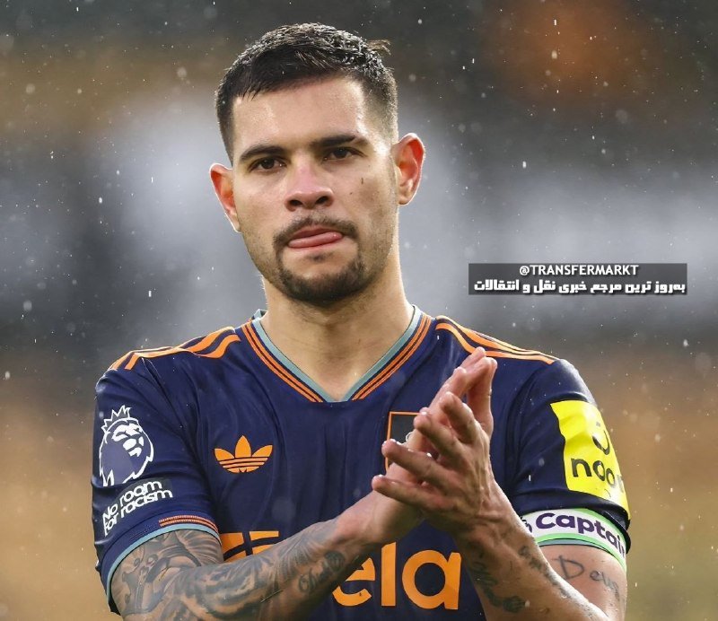 A tattooed soccer player wearing a navy blue Adidas jersey with a captain's armband claps his hands on a rainy field.