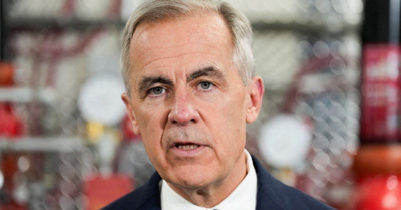 A gray-haired man in a dark suit speaks at a press conference, with a blurred chain-link fence and red signage visible behind him.