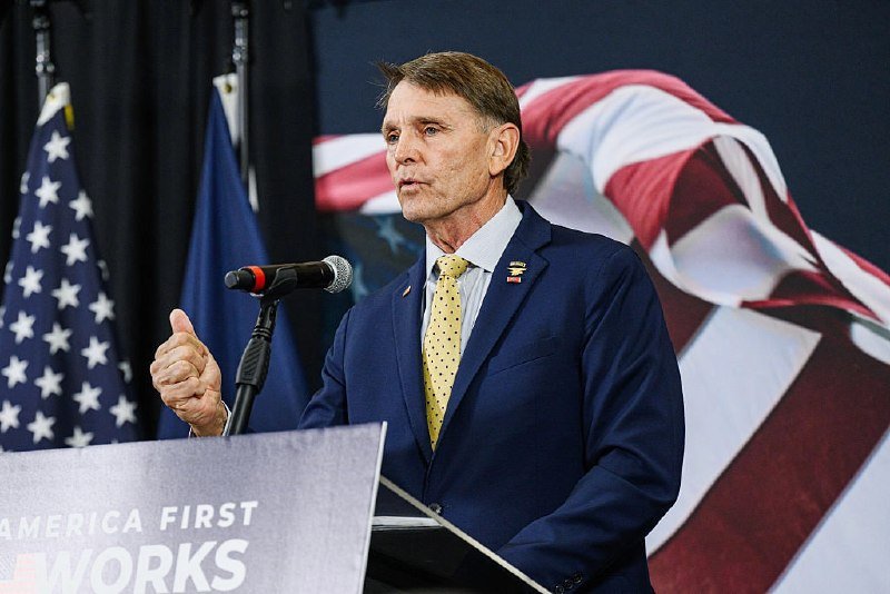 A man in a navy suit and yellow tie speaks at a podium displaying "AMERICA FIRST WORKS," with a U.S. flag and a draped flag visible behind him.
