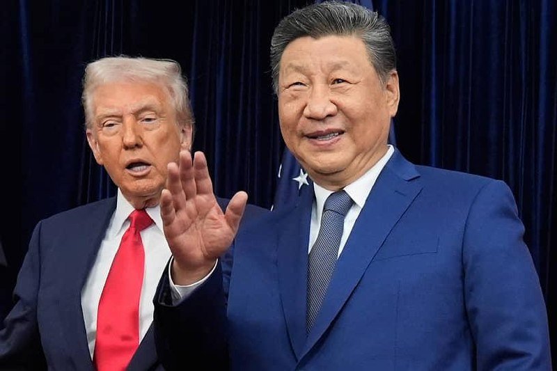 Two men in suits stand side by side against a dark blue curtain backdrop, one with light hair and a red tie gesturing with his hand, the other smiling.
