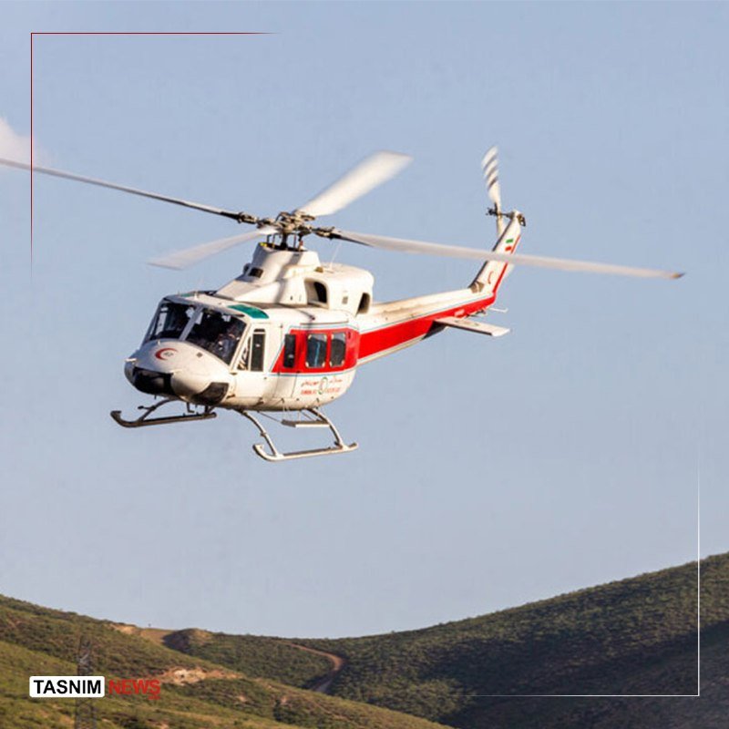 A white and red helicopter flies over a forested hillside beneath a clear sky.
