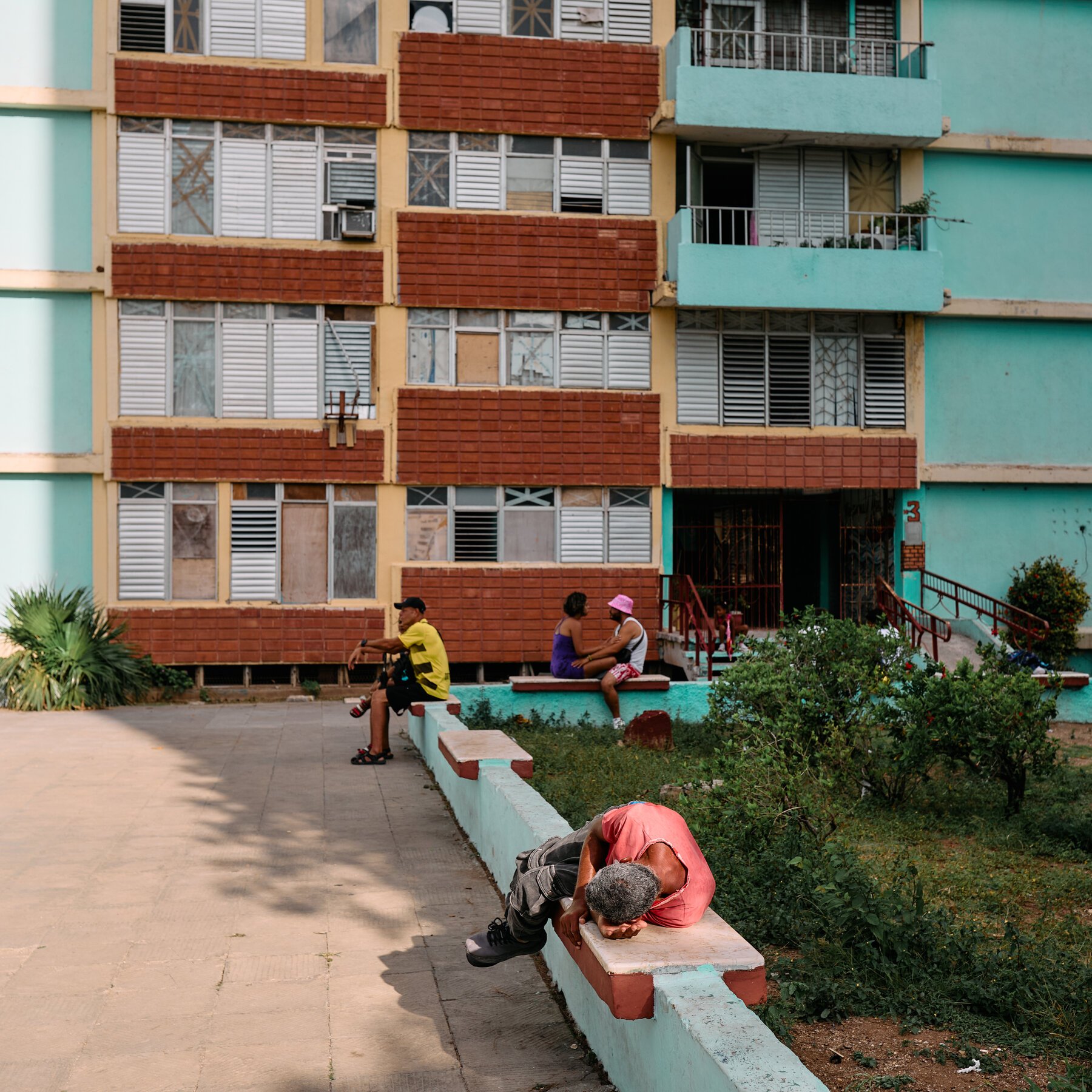 Residents sit and rest on low walls outside a weathered multi-story apartment building with turquoise walls, shuttered windows, and red brick accents.
