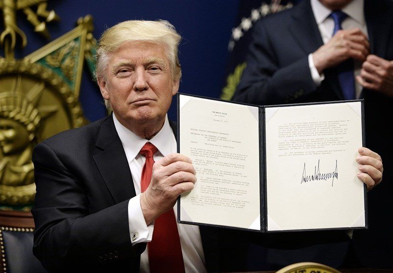 A blond-haired man in a dark suit and red tie holds open a signed document displayed in a black folder, with another suited man standing behind him.
