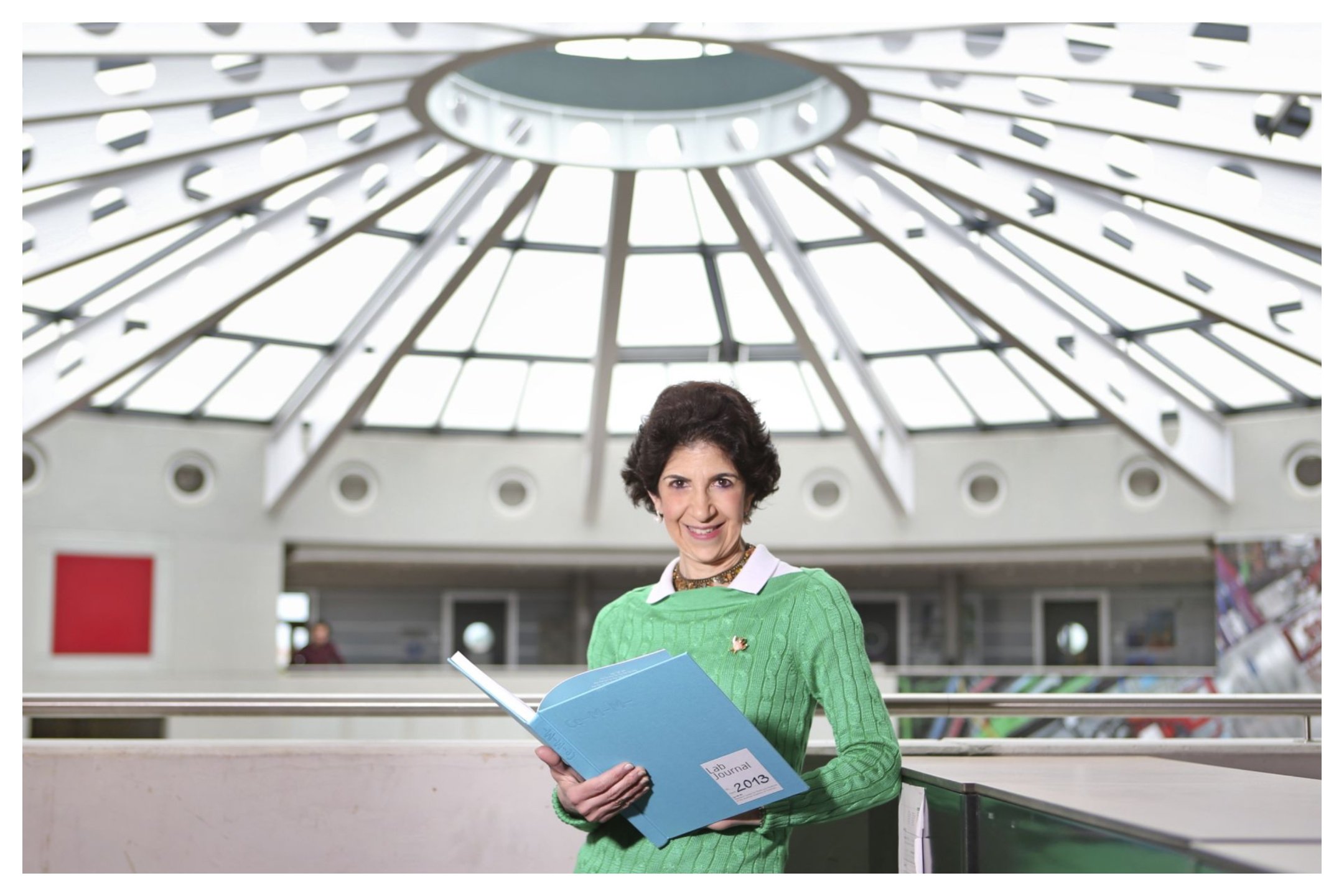 A smiling woman with short dark hair, wearing a green sweater, holds an open blue folder labeled "Lab Journal 2013" inside a modern building with a large domed glass ceiling.