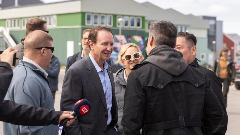 A smiling man in a dark suit speaks with reporters holding a microphone outside a green industrial building.