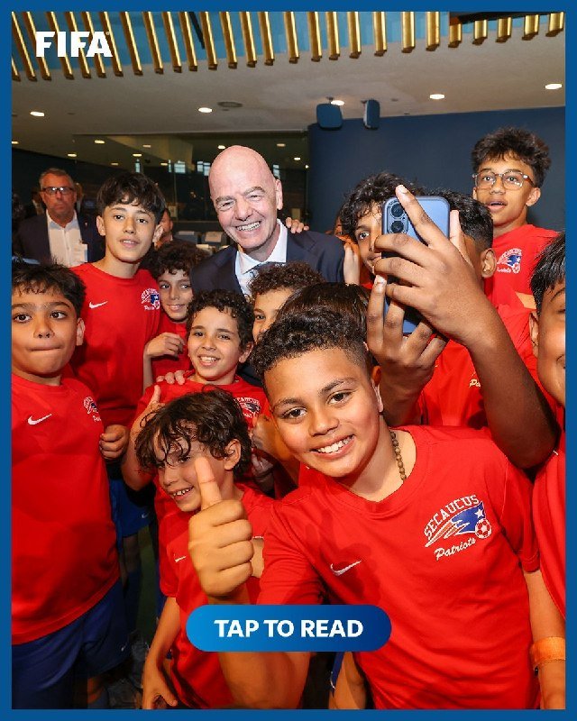 A man in a suit poses for a selfie with a group of boys in red "Sea Lions Patriotas" jerseys, with a "FIFA" logo in the corner.