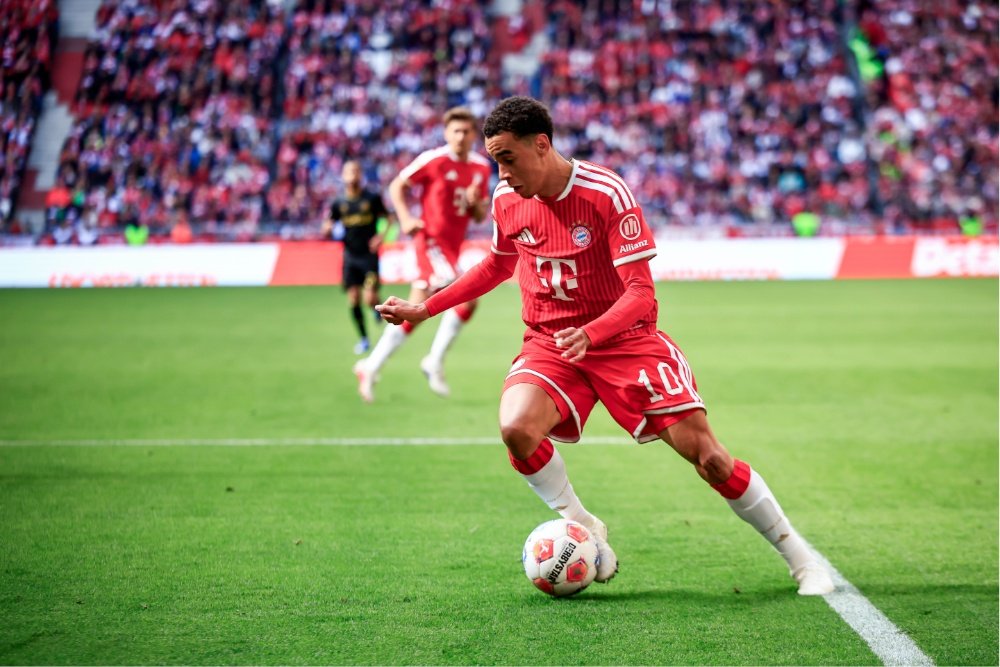 A soccer player in a red Bayern Munich kit controls the ball on a green pitch during a match, with a crowded stadium and a referee visible in the background.