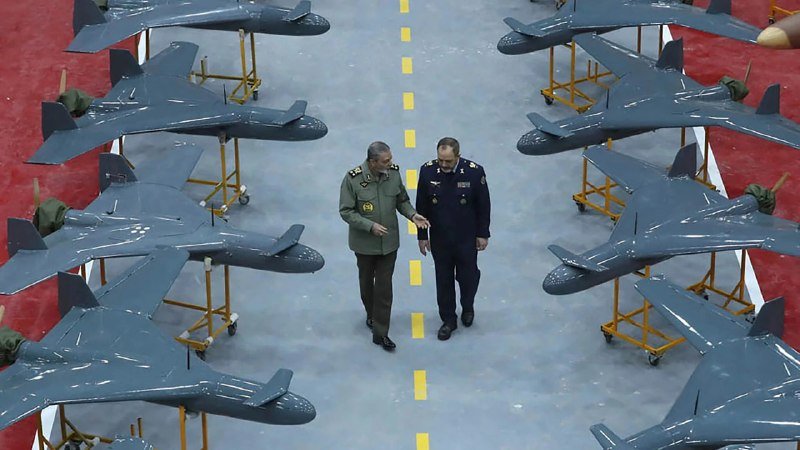 Two uniformed men walk down the center aisle of a hangar lined on both sides with rows of identical blue unmanned aircraft mounted on yellow stands.