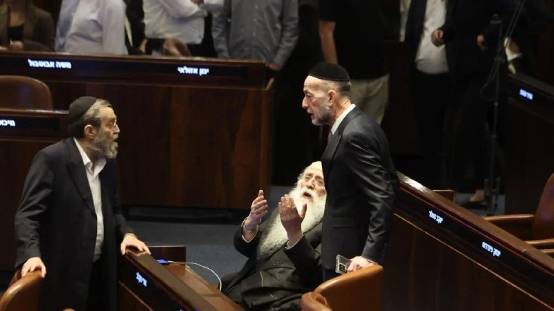 Two bearded men in dark suits and skullcaps converse in a legislative chamber, while a third seated man gestures between them amid Hebrew-labeled desks.