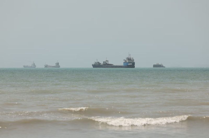 Four cargo ships sit anchored on the horizon of a calm sea under a hazy sky, viewed from a sandy shoreline with gentle waves breaking in the foreground.