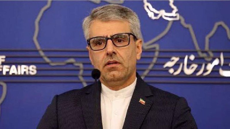 A gray-haired man wearing glasses and a dark suit with an Iranian flag pin speaks at a podium microphone against a blue backdrop displaying a world map outline and partial text reading "FFAIRS."