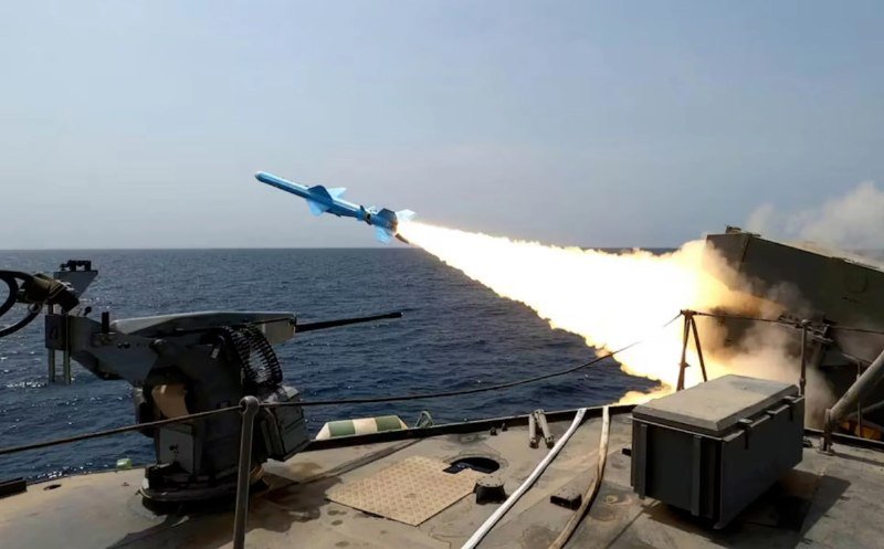 A missile launches from a naval vessel's deck-mounted launcher, propelled by a bright flame trail over the open ocean beneath a hazy sky.