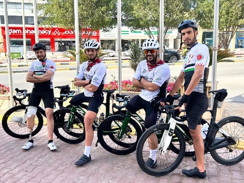 Four helmeted cyclists in matching jerseys stand with their road bikes along a sidewalk in front of storefronts, posing with arms crossed.