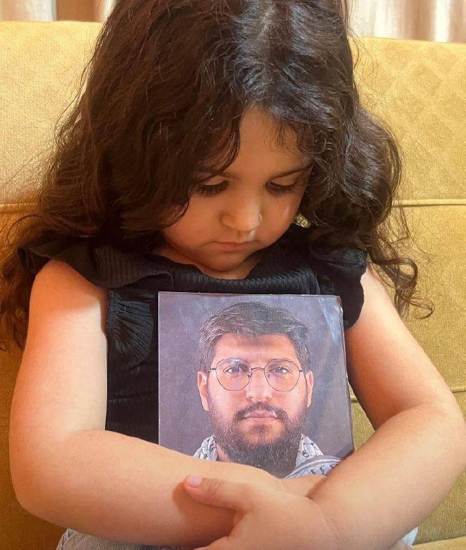 A young girl with curly brown hair looks downward while clutching a printed portrait of a bearded man with glasses against her chest, seated on a yellow sofa.
