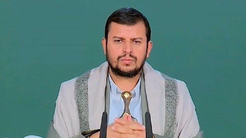 A bearded man wearing a light blue shirt and white shawl stands at microphones, holding a decorative dagger against a green background.
