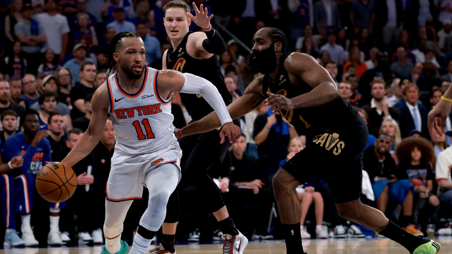 A New York Knicks player wearing number 11 drives with the basketball while being defended by two Cleveland Cavaliers players during an NBA game in front of a crowd.