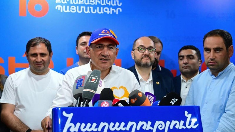 A man in a red and blue cap speaks into multiple microphones at a podium while several men stand behind him against a blue backdrop with Armenian text.